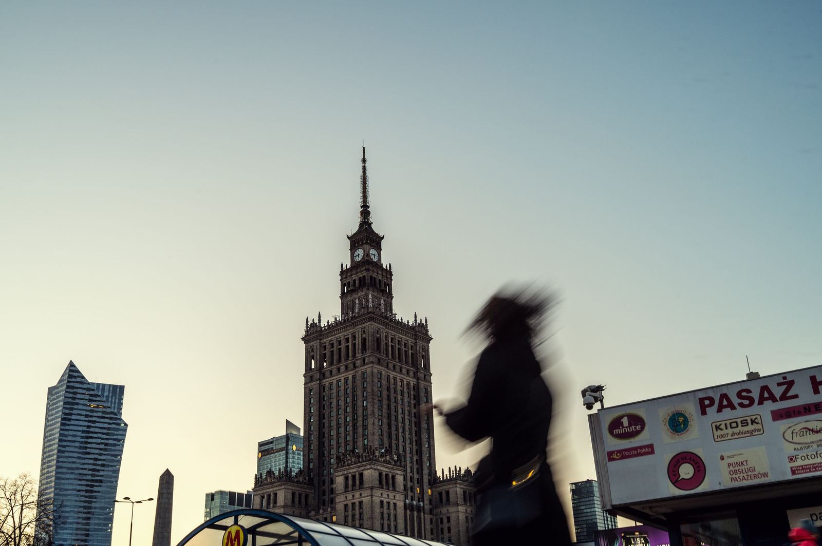Pałac Kultury i Nauki o zmierzchu, z rozmazaną sylwetką przechodnia w ruchu na pierwszym planie i kioskiem z szyldem "Pasaż" przy wejściu do metra.