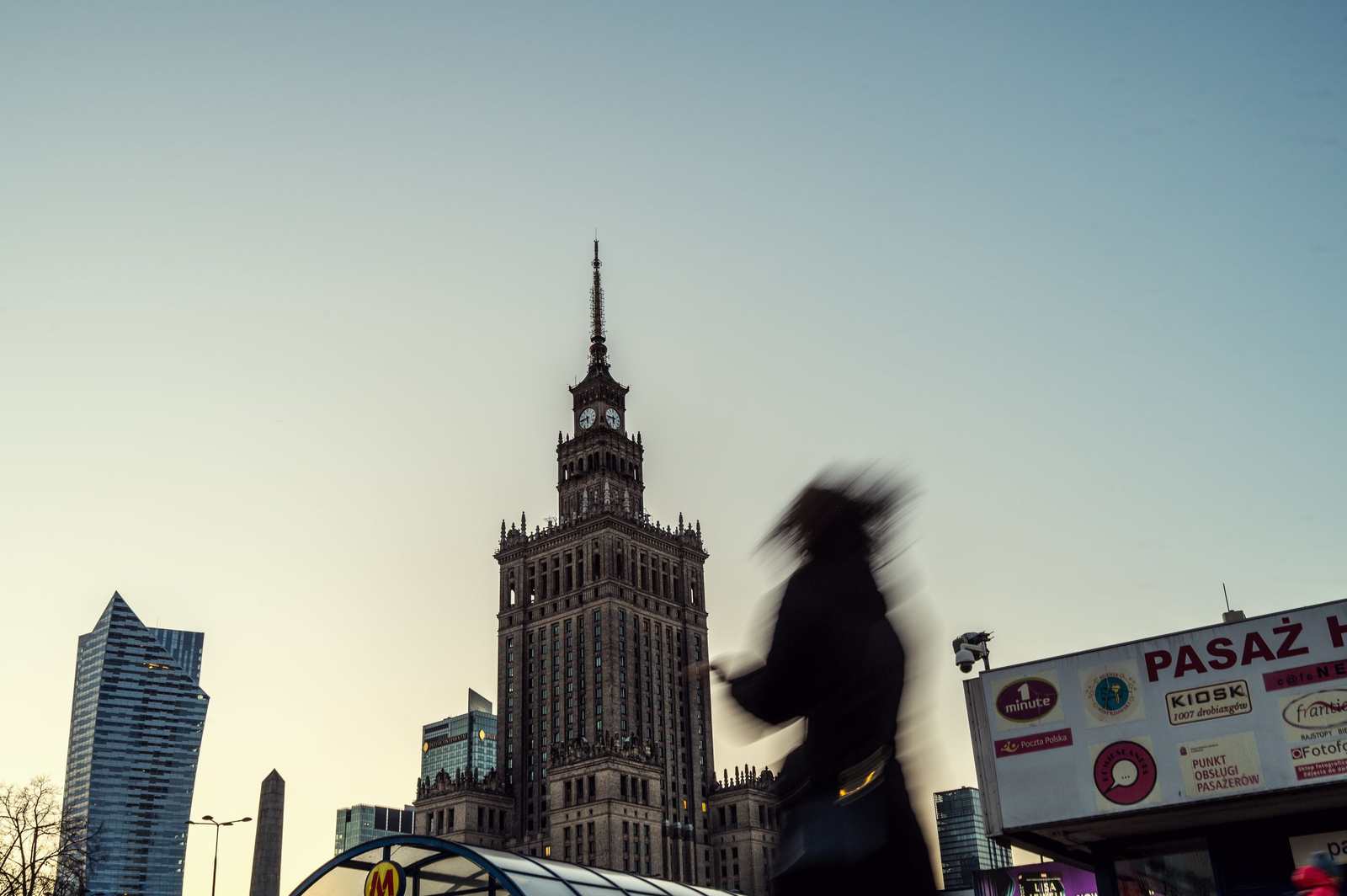 Pałac Kultury i Nauki o zmierzchu, z rozmazaną sylwetką przechodnia w ruchu na pierwszym planie i kioskiem z szyldem "Pasaż" przy wejściu do metra.