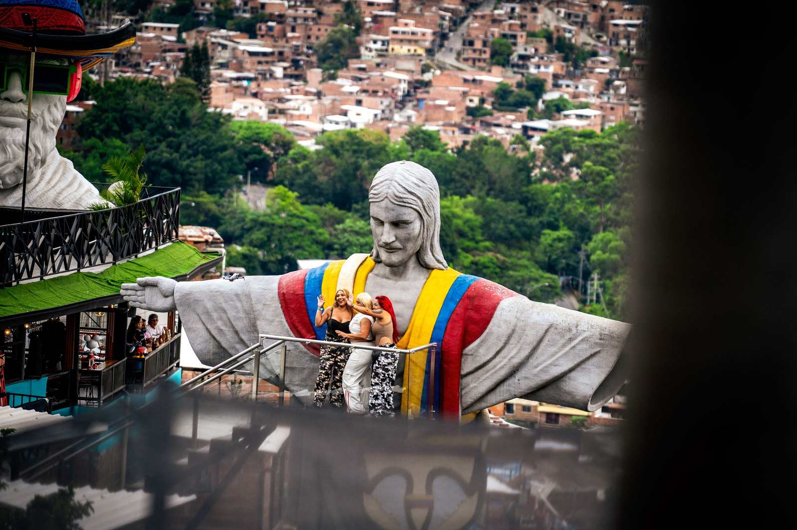 Turystki fotografujące pomnik Chrystusa w barwach Kolumbii na tarasie widokowym w Comunie 13, w tle zabudowa Medellín na zboczu góry.
