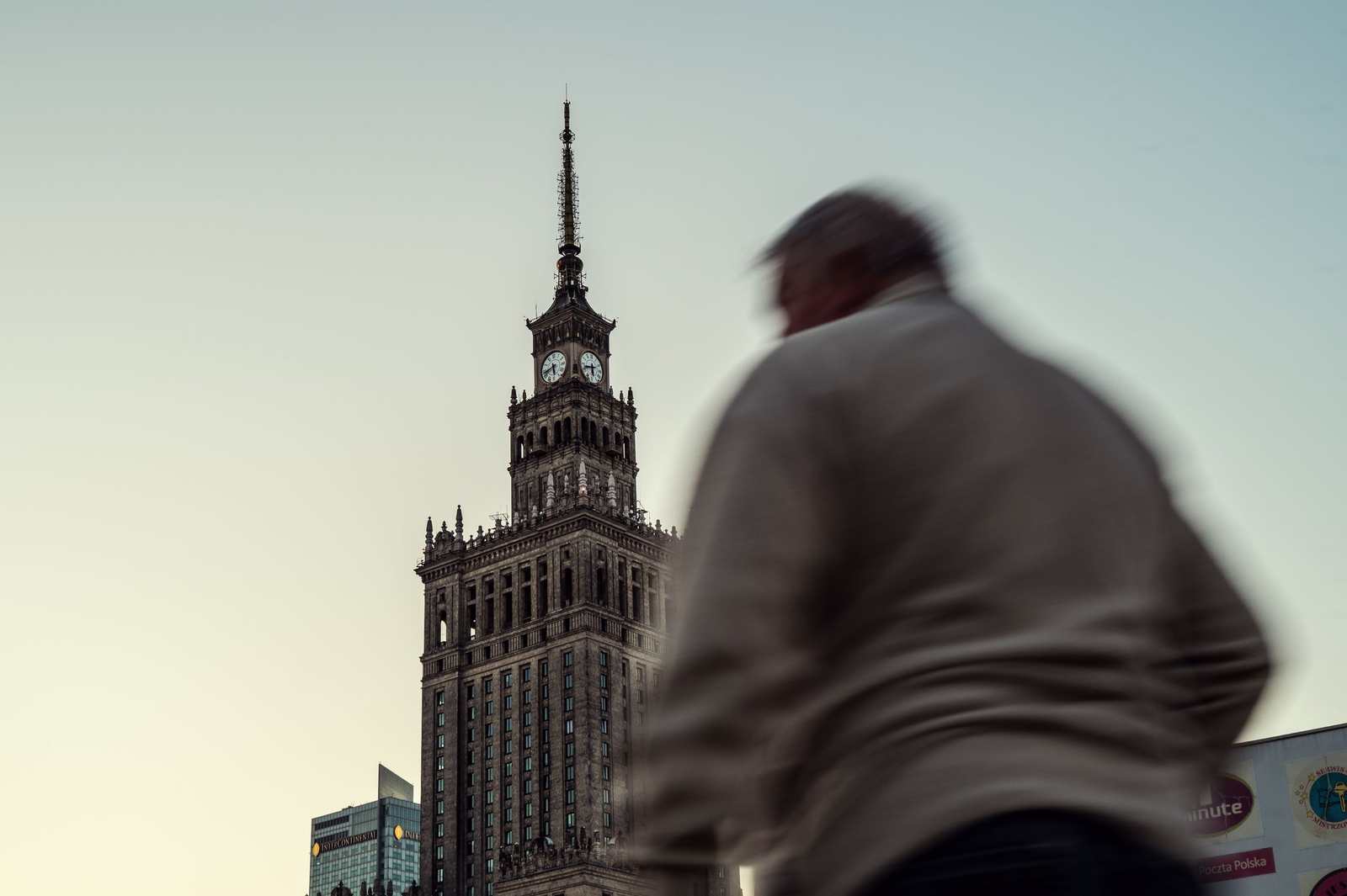 Rozmazana sylwetka starszego mężczyzny w płaszczu na pierwszym planie, za nim wieża zegarowa Pałacu Kultury na tle czystego nieba.