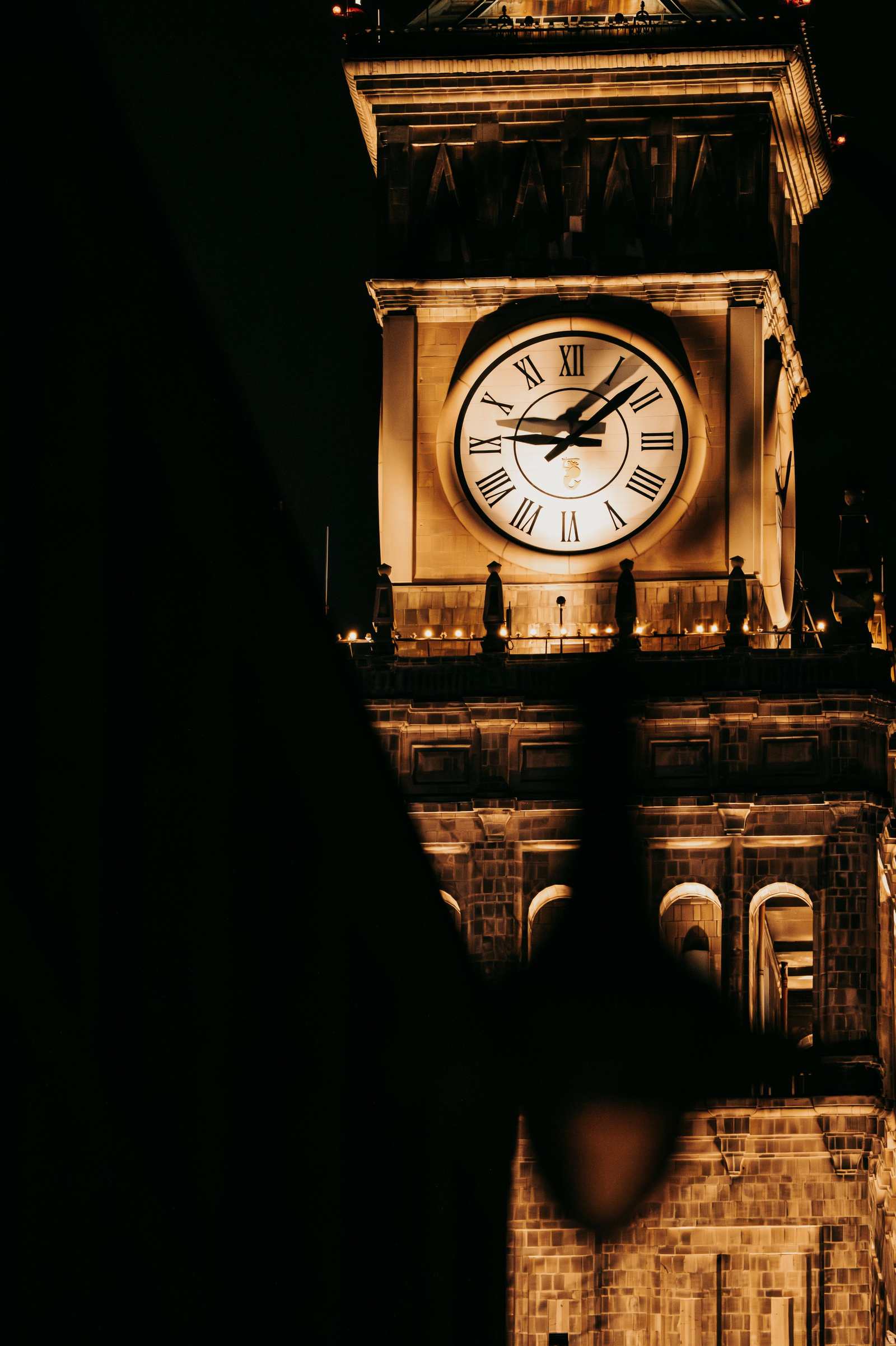 Podświetlony nocą zegar na szczycie wieży Pałacu Kultury, z ciemnym kształtem w rogu kadru zasłaniającym część iglicy.