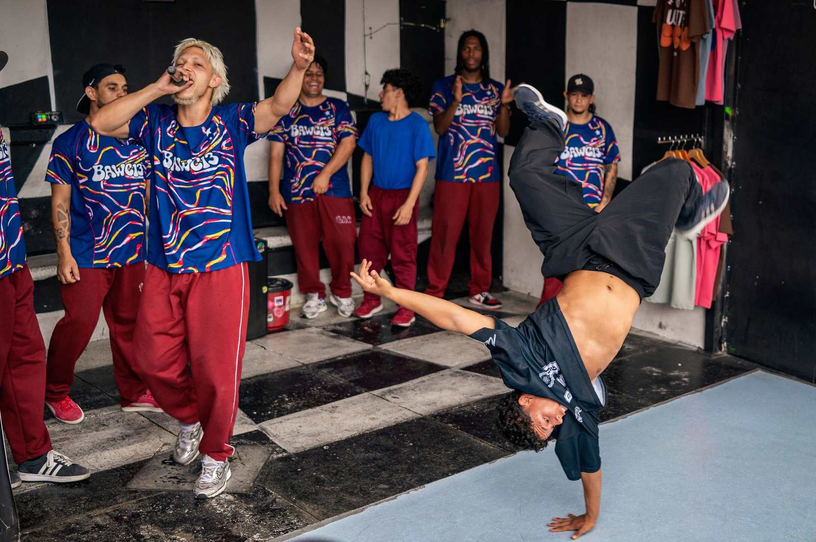 Tancerz breakdance w akrobatycznym wygięciu przed grupą w koszulkach zespołu "BAWC13".