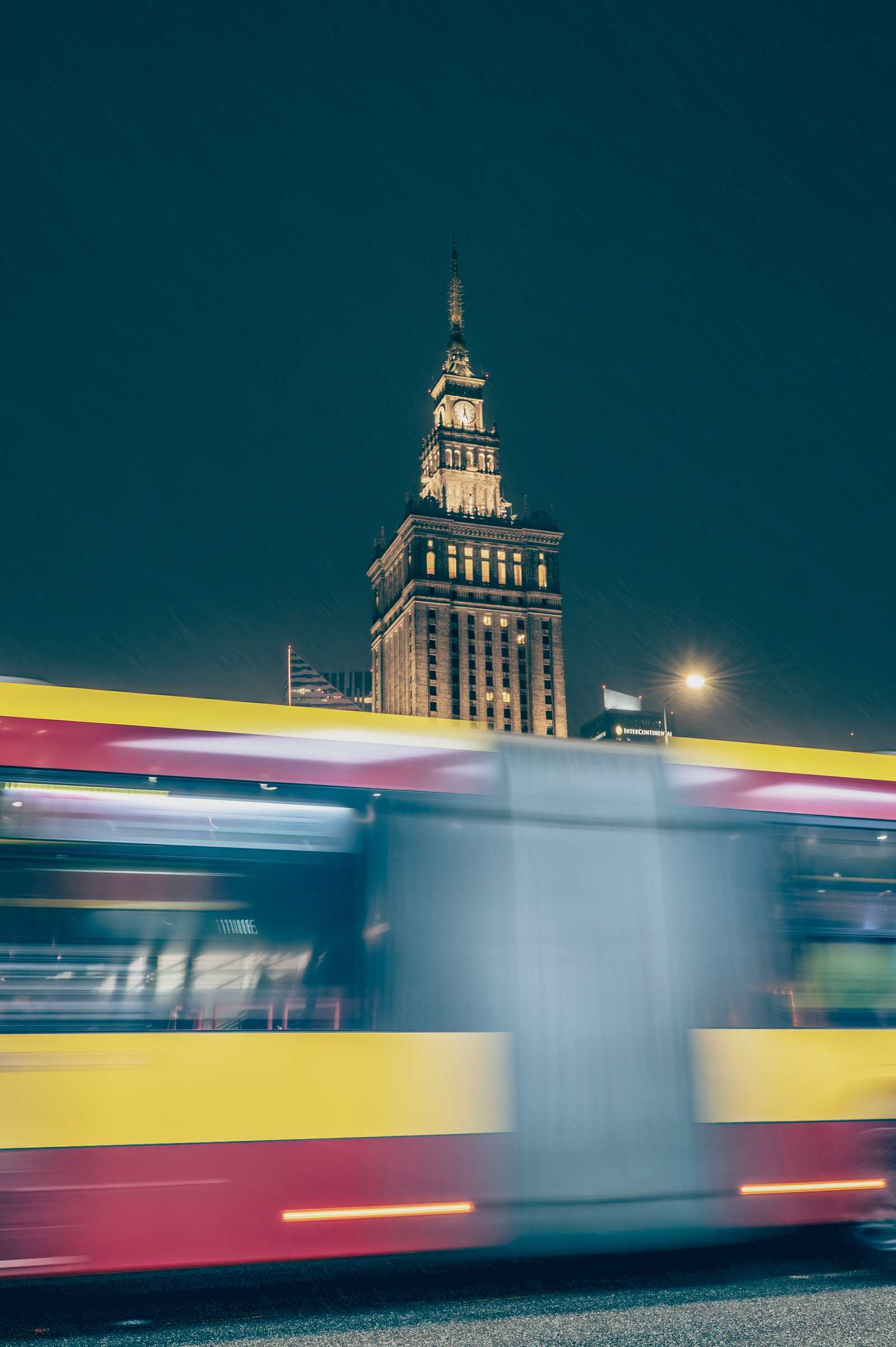 Rozmazany w ruchu autobus miejski przejeżdżający nocą na tle podświetlonego Pałacu Kultury, w deszczu.