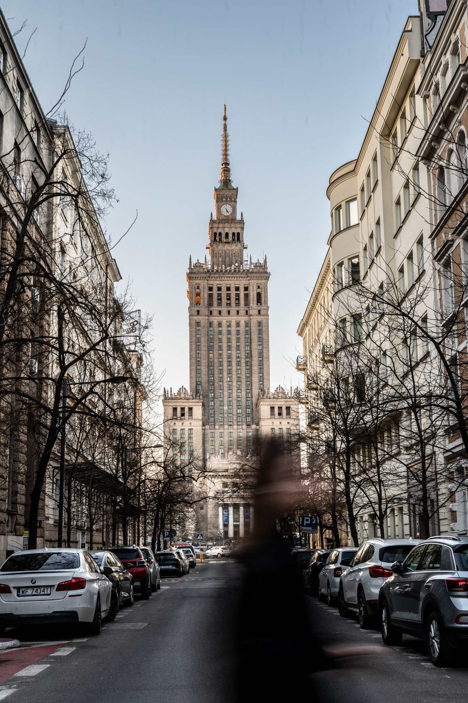 Widok w dół warszawskiej ulicy obudowanej kamienicami i zaparkowanymi samochodami, z Pałacem Kultury na wprost w złotym świetle.
