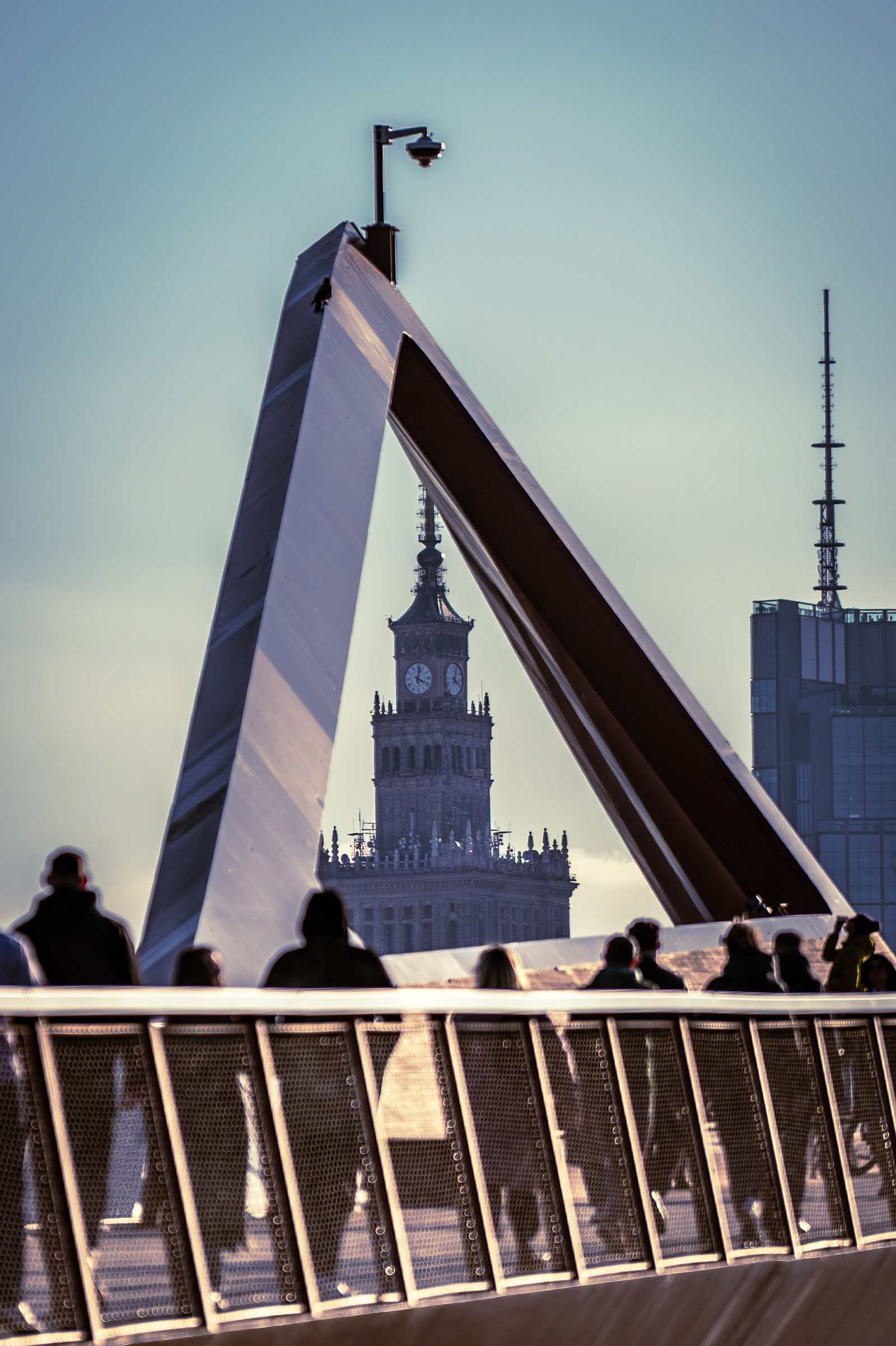 Wieża Pałacu Kultury widziana z mostu pieszo-rowerowego, wpisana między stalowe belki konstrukcji i sylwetki przechodniów.