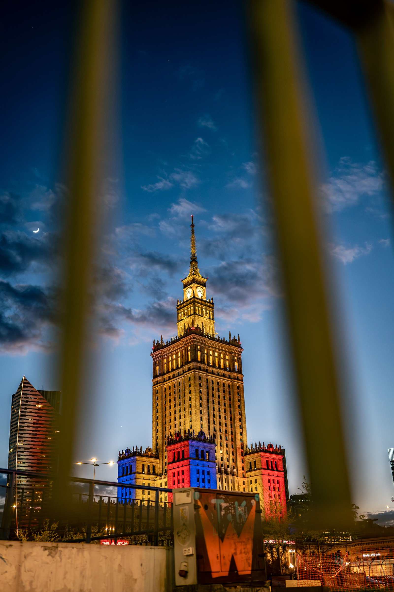 Pałac Kultury nocą podświetlony na czerwono i niebiesko, kadr przez żółte pręty barierki, w rogu sierp księżyca.