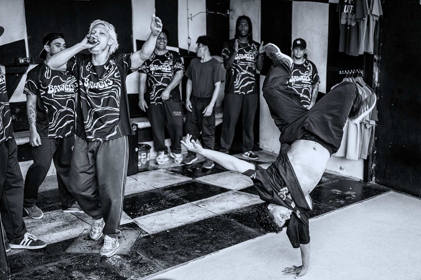 Tancerz breakdance stoi na jednej ręce do góry nogami, a ekipa w niebieskich koszulkach z napisem BAWC13 patrzy; z boku konferansjer z mikrofonem.