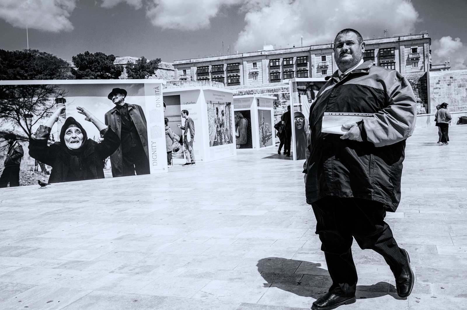 Mężczyzna w ciemnej kurtce z plikiem kartek pod pachą przechodzi obok plenerowej wystawy fotografii na kamiennym placu w Valletcie.