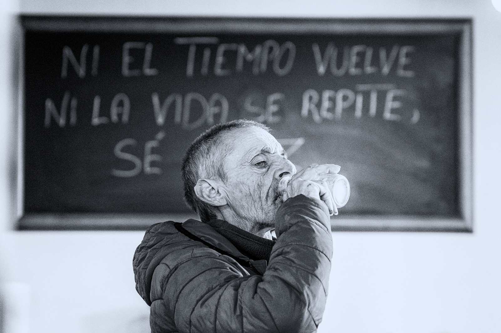Mężczyzna w czerwonej kurtce pije ze szklanki pod tablicą z kredowym napisem NI EL TIEMPO VUELVE NI LA VIDA SE REPITE.