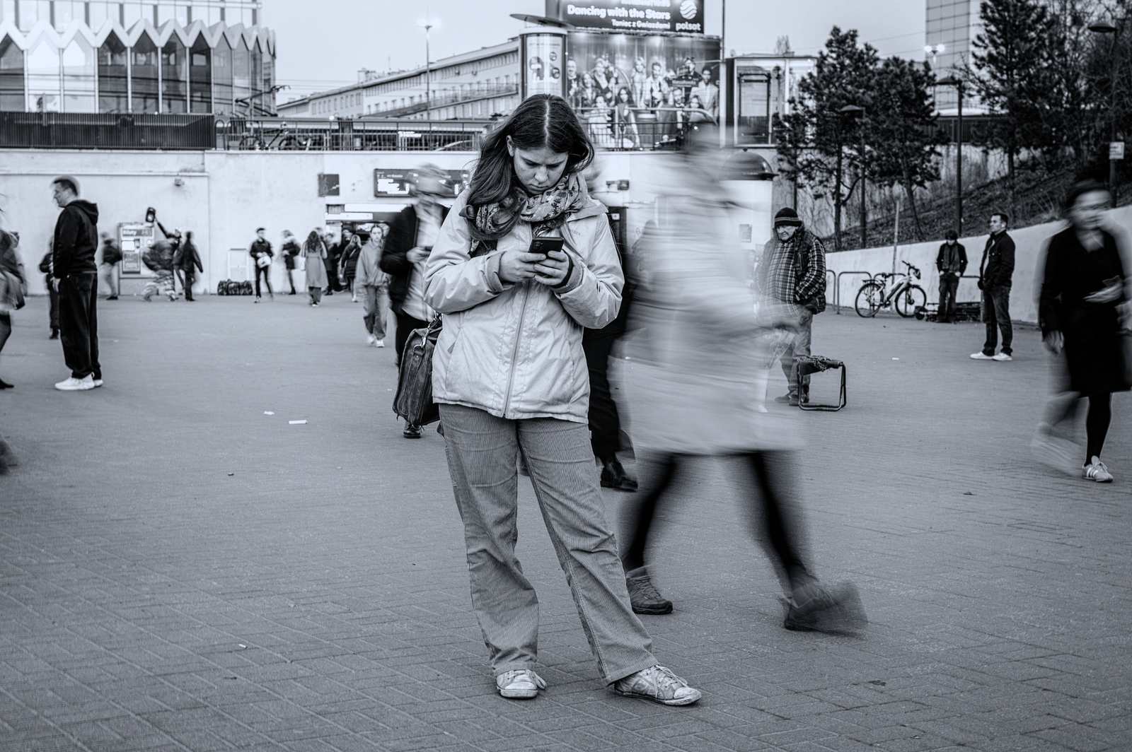 Kobieta w jasnej kurtce stoi nieruchomo z telefonem w dłoniach, a rozmazany ruchem przechodzień mija ją przed wejściem do metra.