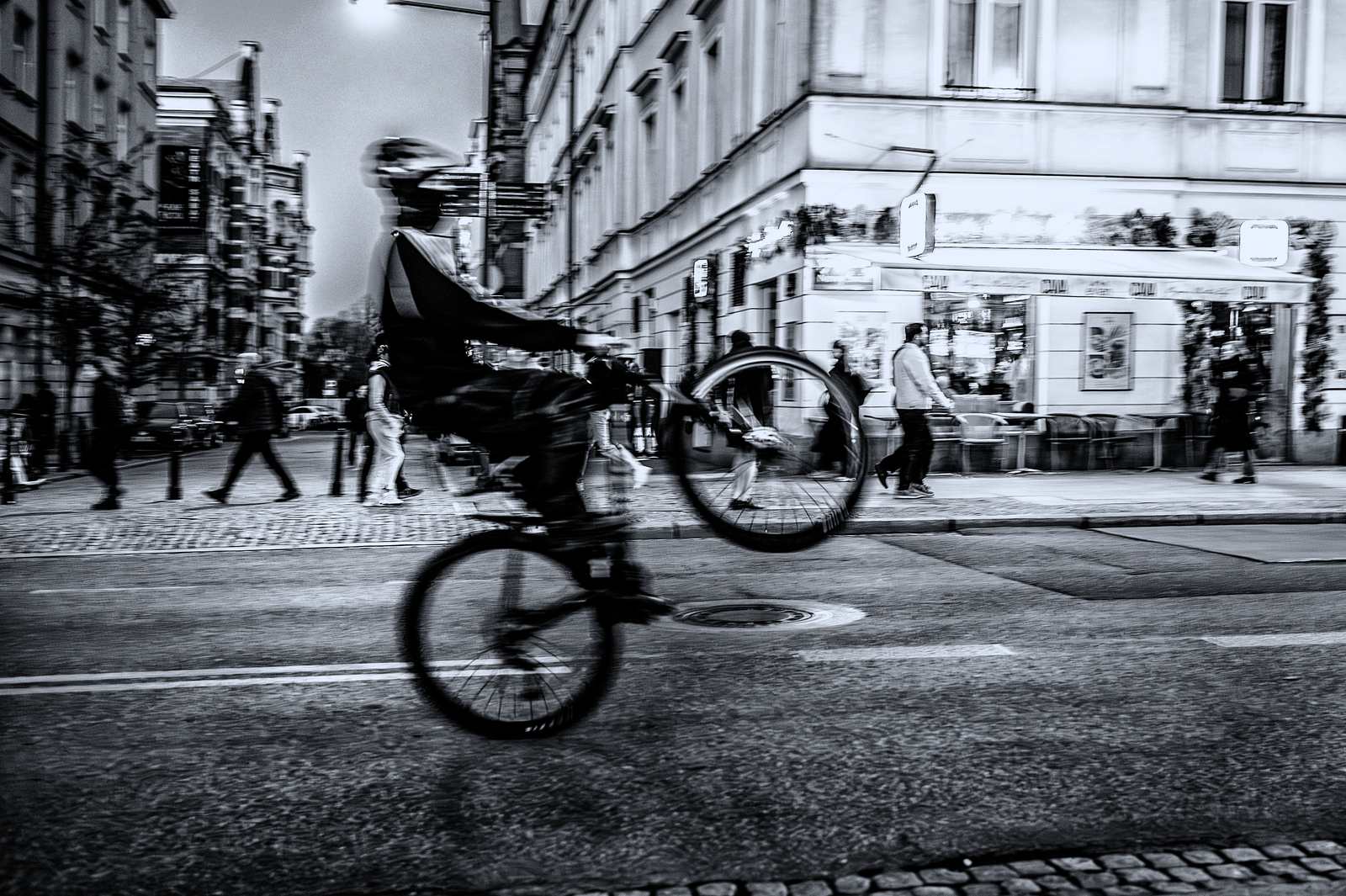 Rozmyty ruchem rowerzysta przejeżdża wieczorem przez skrzyżowanie; w tle rozświetlona witryna kawiarni i przechodnie.